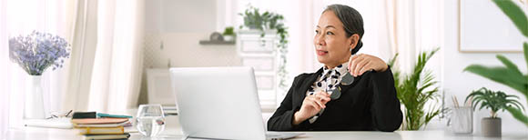 Decorative. A dignified female executive in professional dress seated in her office surrounded by potted plants. She stares out the window contemplatively while holding a pair of eyeglasses with both hands.