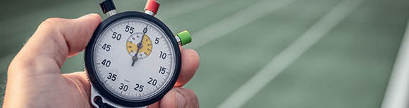 On the left, a hand holds an analog stopwatch. In the background, there is a track for foot races with multiple lanes.