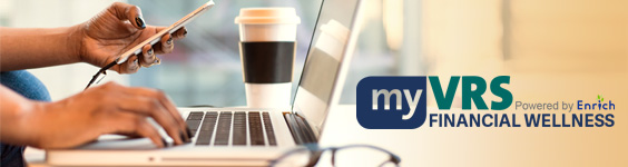 A pair of hands holding a cellphone in the left hand and typing on a laptop computer with the right. A coffee cup with a lide is to the left of the laptop on a table. A pair of eyeglasses rests on the table in the foreground. On the right side of the image past the laptop, the myVRS Financial Wellness logo is superimposed over part of the table and wall.