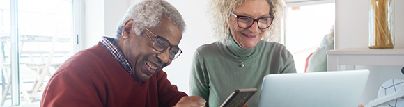 A retired couple smiling while looking at the screens of a laptop computer and a handheld tablet.