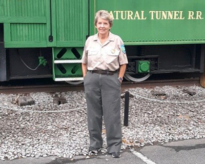 a full length photo of Park Ranger Saundra Tomlinson standing in front of a rail car that says Natural Tunnel R. R. on the side