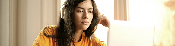 A young woman seated at a desk, wearing headphones, staring at a laptop computer.