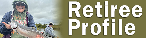 Denise Grant stands in a boat on the water holding a freshly caught salmon. A man in the background looks on.