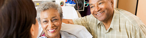 An older black woman in a hospital bed and an older black man next to her bed smile broadly at a  woman across from them.