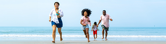 An African family of four wearing summer clothing run along a sunny beach toward the viewer.