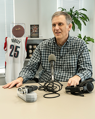 Rohn Brown seated before a microphone on a table. A collection of sports memorabilia is behind him.